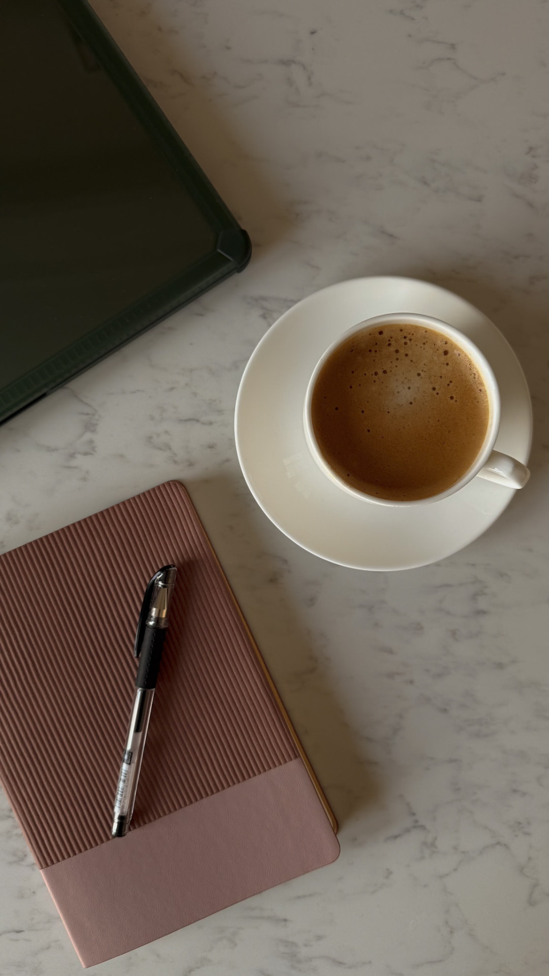 Overhead flat lay of a coffee cup, rose notebook, pen and tablet on a marble surface