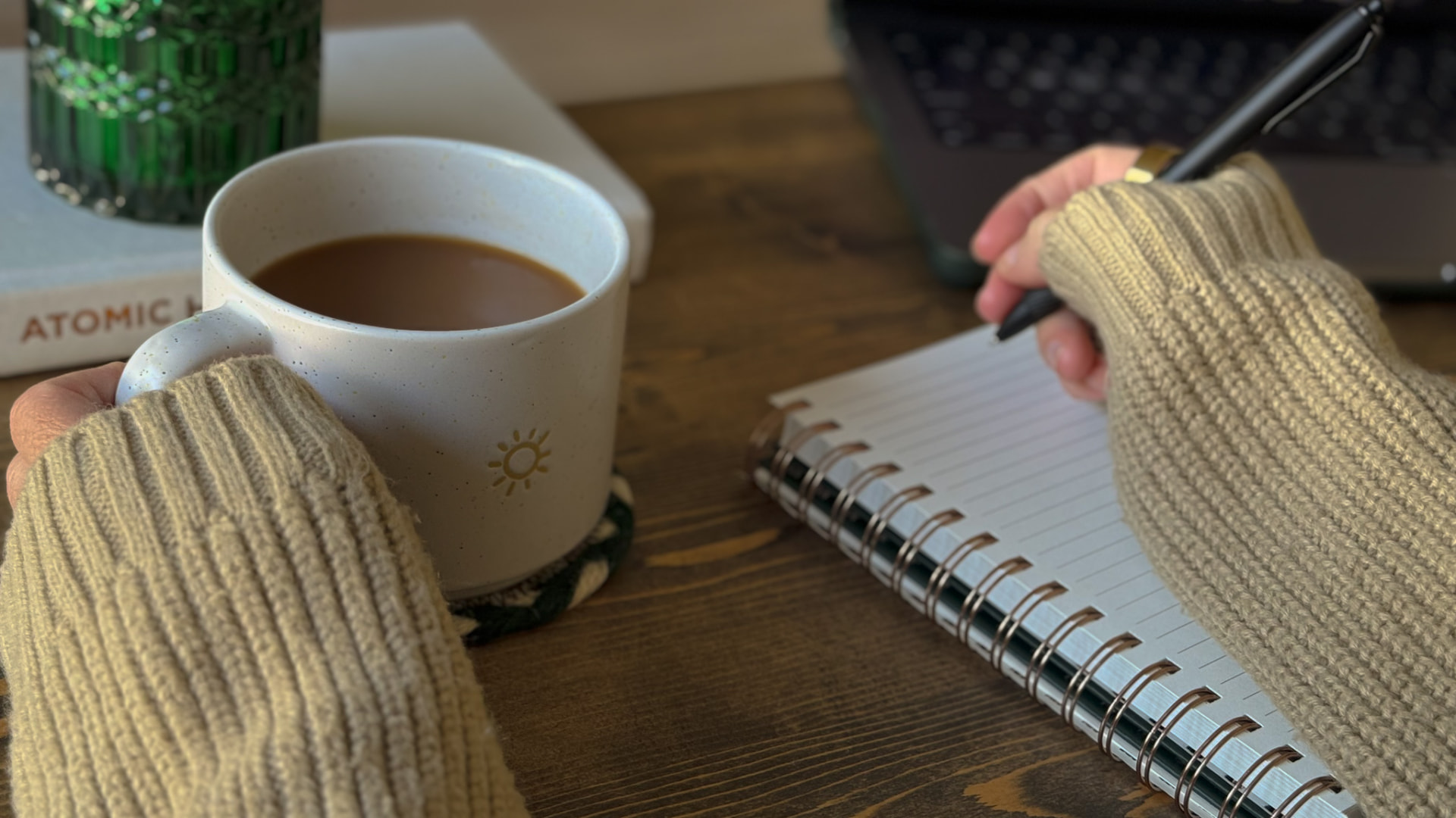 Hands holding a coffee mug and writing in a spiral notebook on a warm wooden table