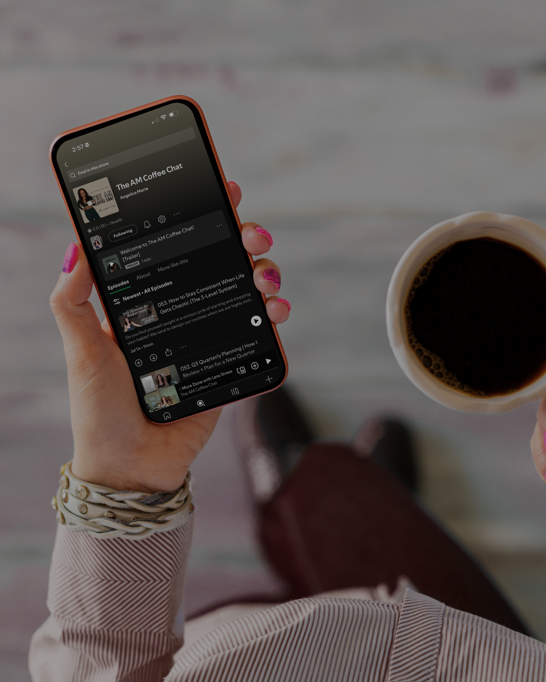 Hand holding a phone playing The AM Coffee Chat podcast next to a cup of coffee
