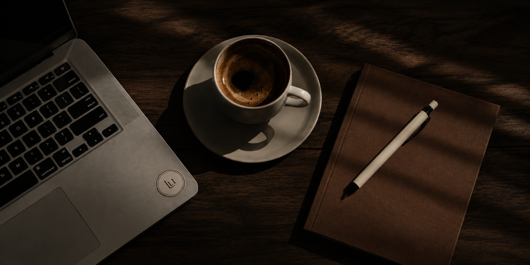 Overhead flat lay of a silver laptop, ceramic espresso cup, brown journal and cream pen on a dark wood table